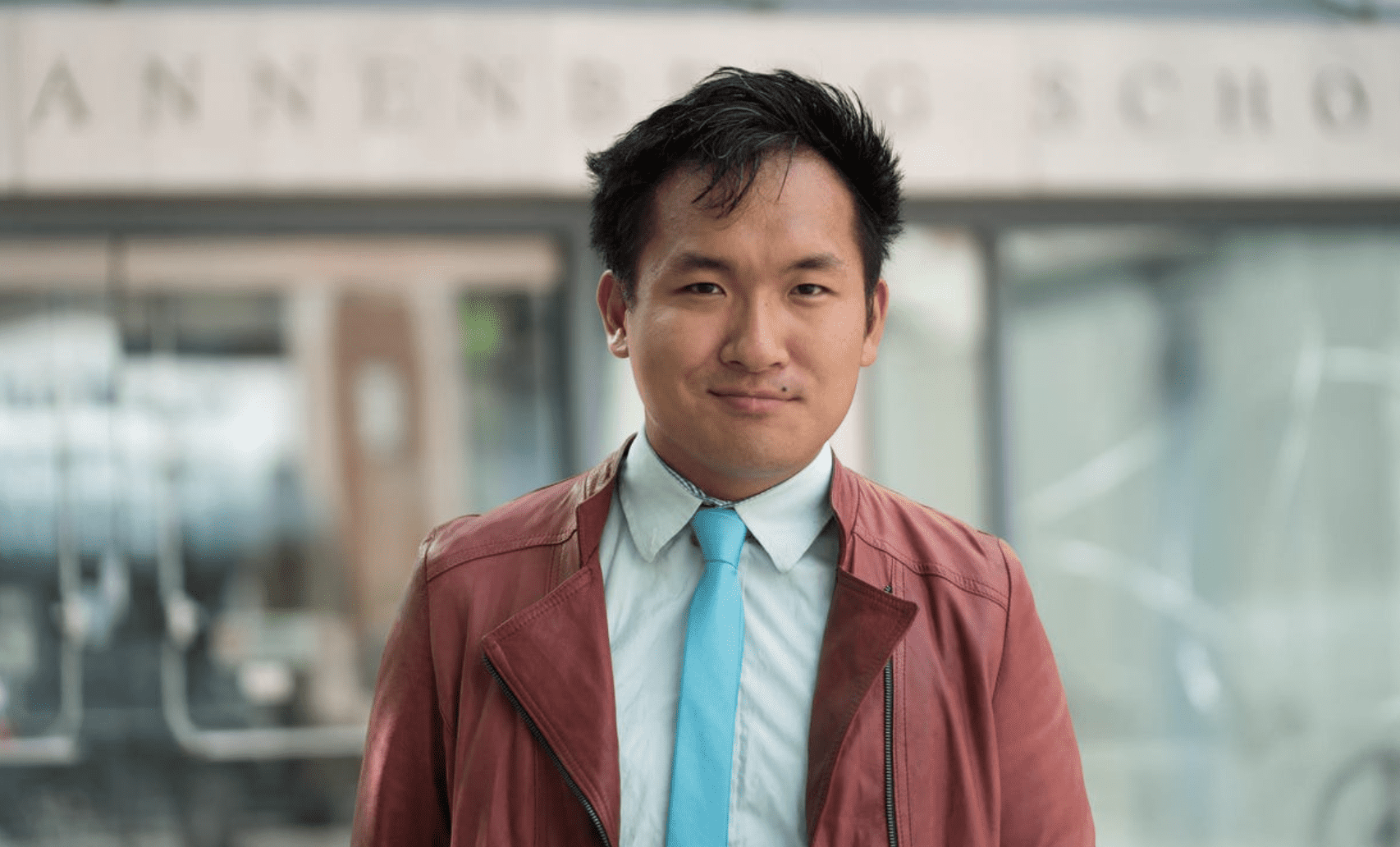 A young Asian man in a casual outfit standing outdoors, smiling confidently, with a blurred cityscape background. Ideal for content about urban lifestyle, diversity, or modern urban fashion.