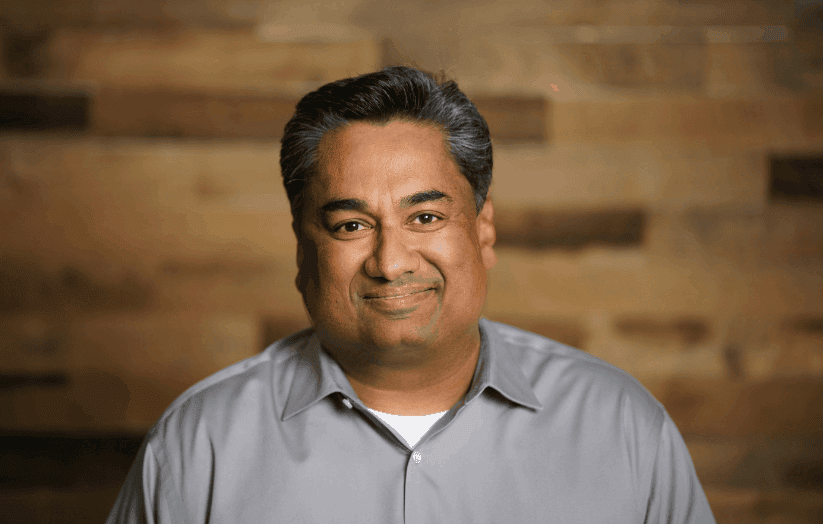 Business professional portrait, smiling man in gray shirt, studio background with warm wooden paneling, confident expressions, professional headshot for corporate or marketing use.