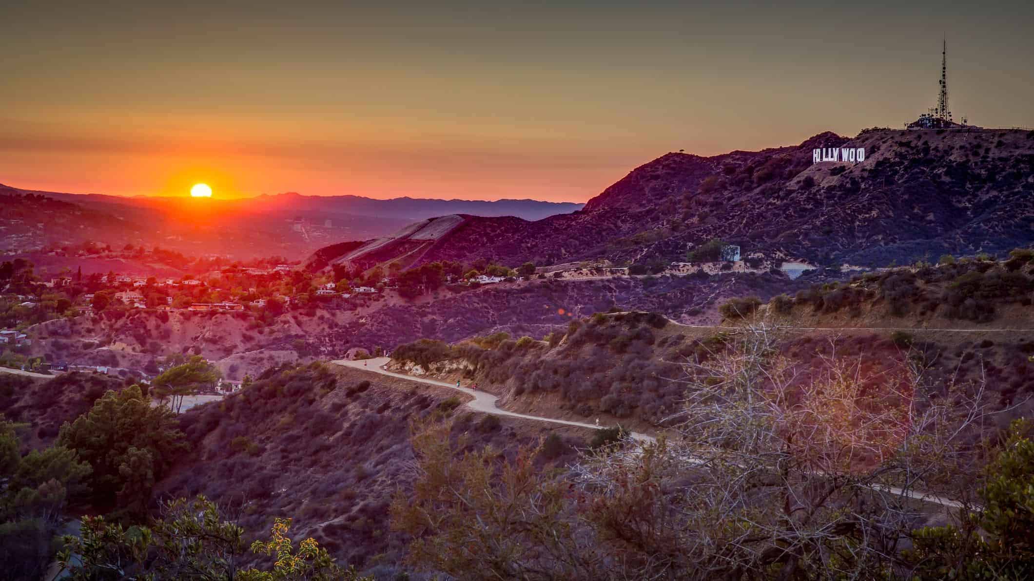 Vibrant sunset over the Hollywood Hills with the iconic Hollywood Sign illuminated, showcasing scenic landscape, winding road, and lush vegetation under warm sky.