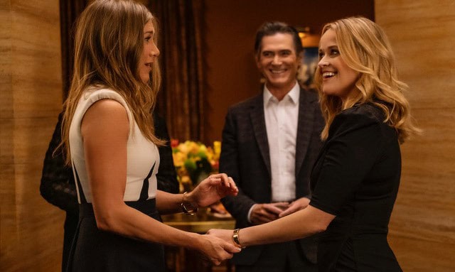 A woman and a man holding hands and smiling at each other during a social event, with another smiling man in the background, showcasing a warm, friendly atmosphere and connection.