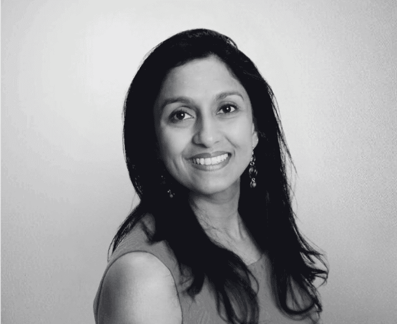 A woman with long dark hair smiling at the camera, wearing earrings and a sleeveless top, in a professional portrait setting.