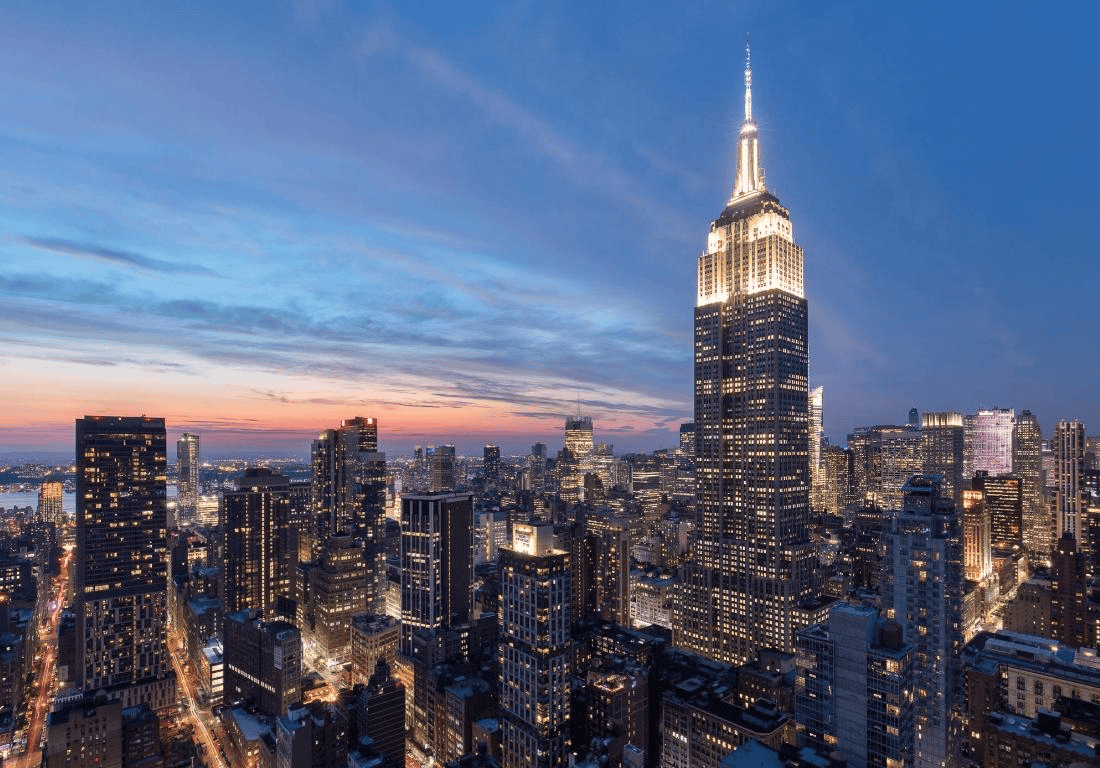 Night view of the Empire State Building illuminated in New York City during sunset, with surrounding skyscrapers and a colorful sky.